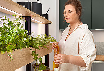 Frau in einer modernen Küche kümmert sich um ihren Indoor-Kräutergarten, der mit natürlichem Licht durchflutet ist und einen gesunden, nachhaltigen Lebensstil zu Hause präsentiert.