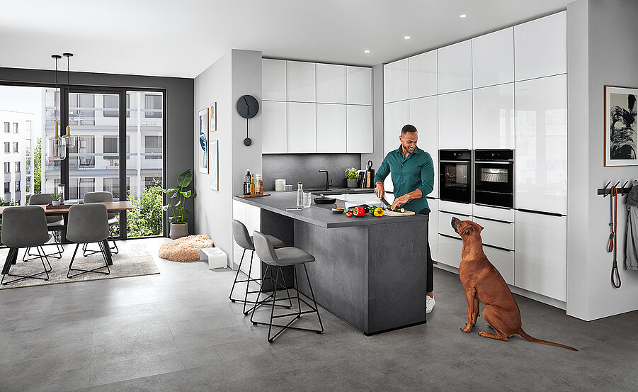 Ein modernes Kücheninterieur mit einem Mann, der Essen auf der Arbeitsplatte zubereitet, während ein Hund in der Nähe sitzt, zeigt elegante Geräte und einen gut beleuchteten Essbereich.
