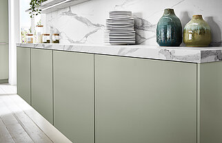 Moderne Küche mit klaren Linien, die Salbeigrüne Schränke, Marmorarbeitsplatten und eine minimalistische Anordnung von Keramikgeschirr und Vasen zeigt.