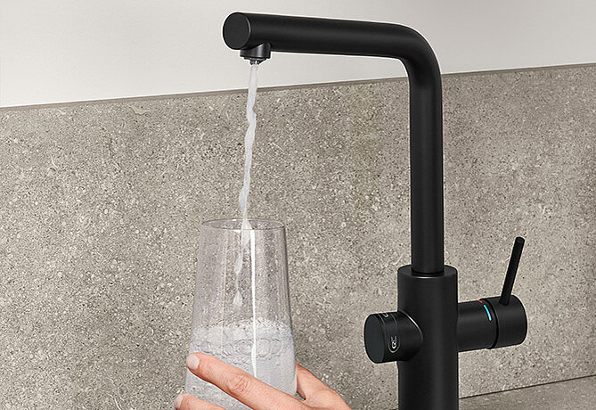 Matt-schwarzer moderner Küchenarmatur mit separatem Wasserspender für gefiltertes Wasser, der ein klares Glas füllt, das von einer menschlichen Hand gegen einen grauen Spritzschutz gehalten wird.