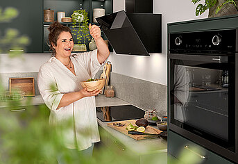 Eine lächelnde Person würzt ihr Essen in einer modernen Küche mit stilvollen schwarzen Geräten und grüner Pflanzen im Hintergrund.