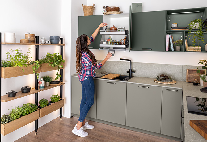 Frau in einer modernen Küche mit grünen Schränken, die nach Gegenständen greift, umgeben von frischen Kräutern und Holzregalen, die einen umweltfreundlichen Lebensstil darstellen.