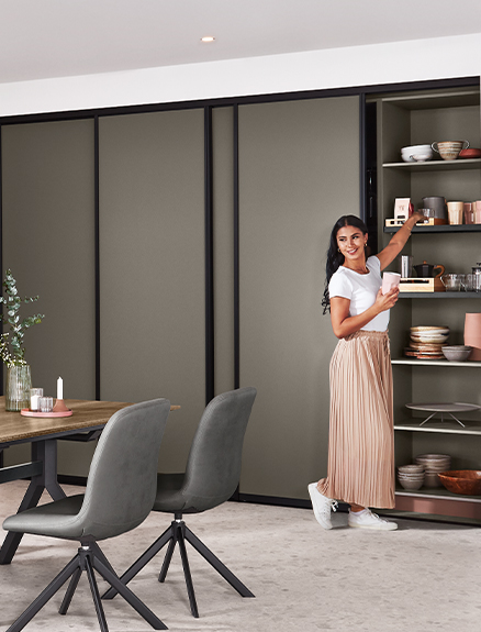 Ein zeitgemäßes Esszimmer mit einer lächelnden Frau neben einem offenen Regal, mit stilvollen Möbeln und einem minimalistischen Farbschema.