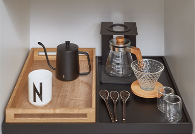 Eine elegant arrangierte Kaffee-Station mit einem schwarzen Wasserkocher, einer Mühle, Gläsern und einzigartigen Löffeln auf einem schicken Holztablett, das modernes Küchengeschirr-Design präsentiert.
