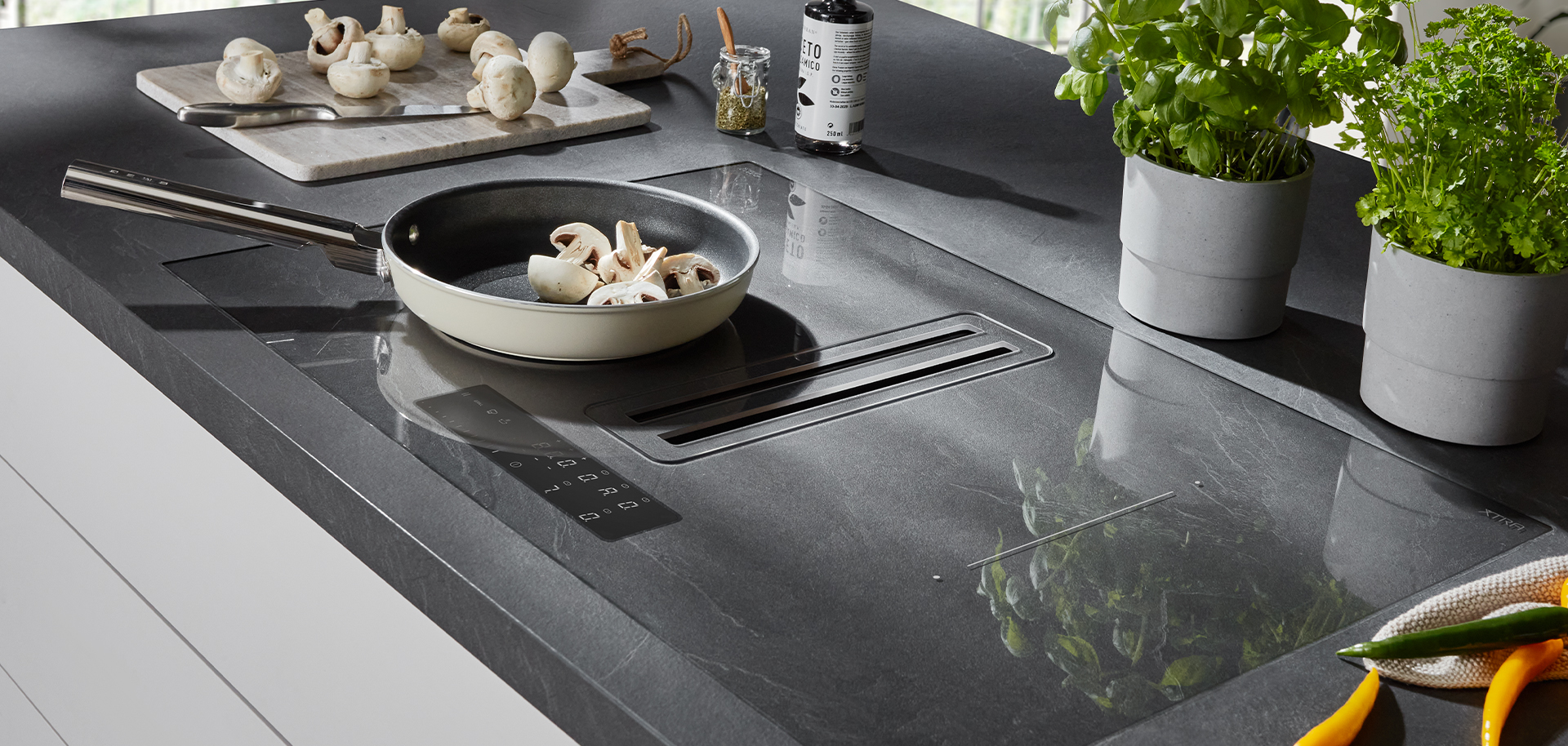Eine moderne Küchenarbeitsplatte mit einem Induktionskochfeld mit einer Pfanne voller Pilze, umgeben von frischen Kräutern und schlichter, minimalistischer Dekoration.
