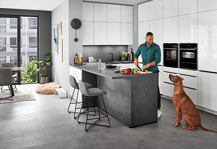 Eine moderne Küche mit klaren Linien, auf der eine Person Essen auf einer eleganten Kücheninsel zubereitet, während ein Hund aufmerksam zuschaut.